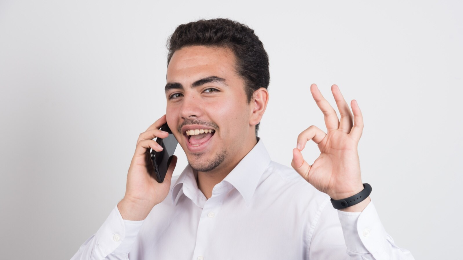 A cheerful man gives an "OK" hand gesture while speaking on the phone, wearing a white shirt. This scene portrays satisfaction with a communication solution, possibly related to effective VoIP call recording or Teams call recording features for compliance.