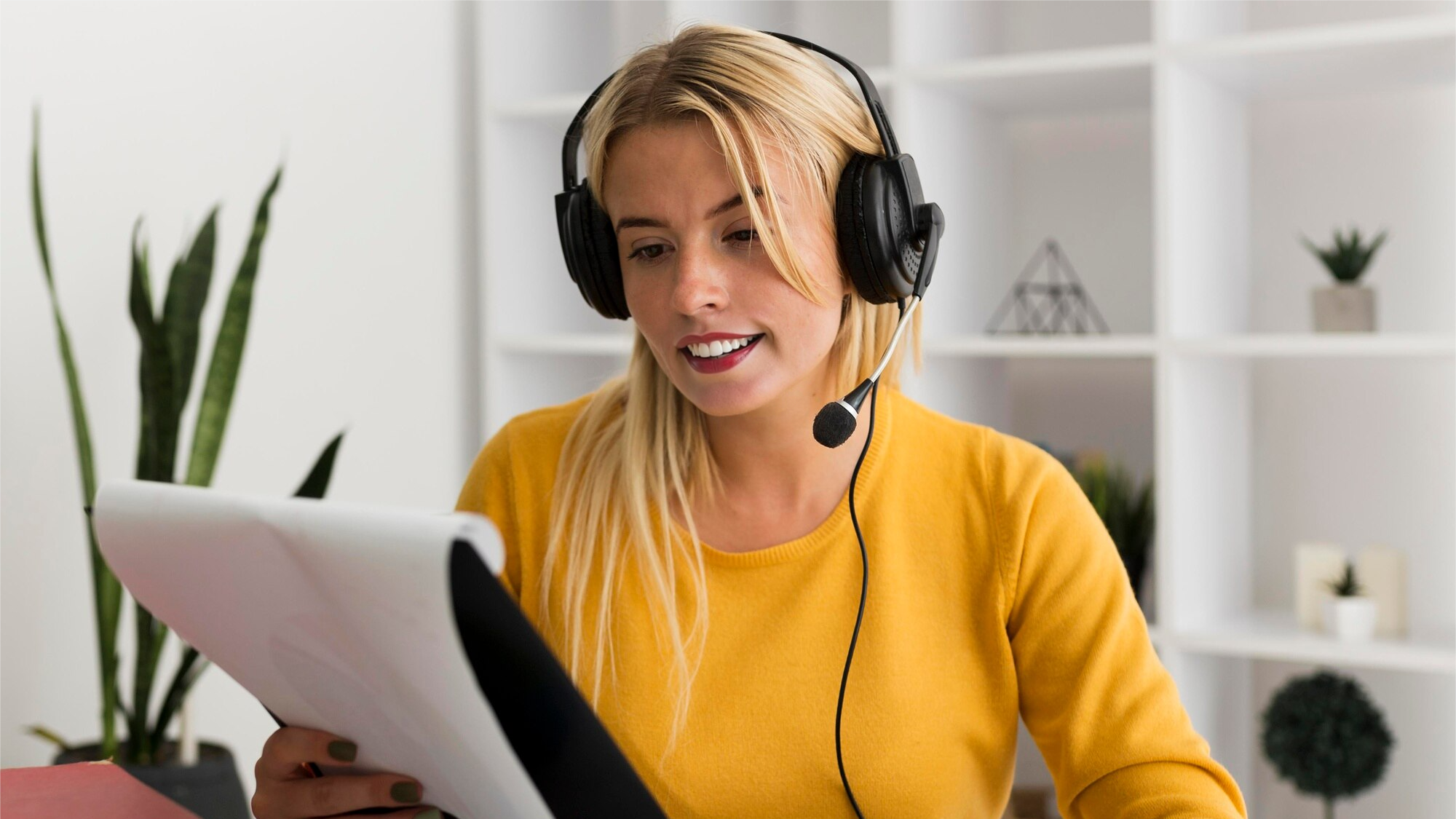 Young woman with a headset smiling and speaking while holding papers during a virtual meeting, possibly engaged in a Teams call with active phone recording for documentation or training purposes.