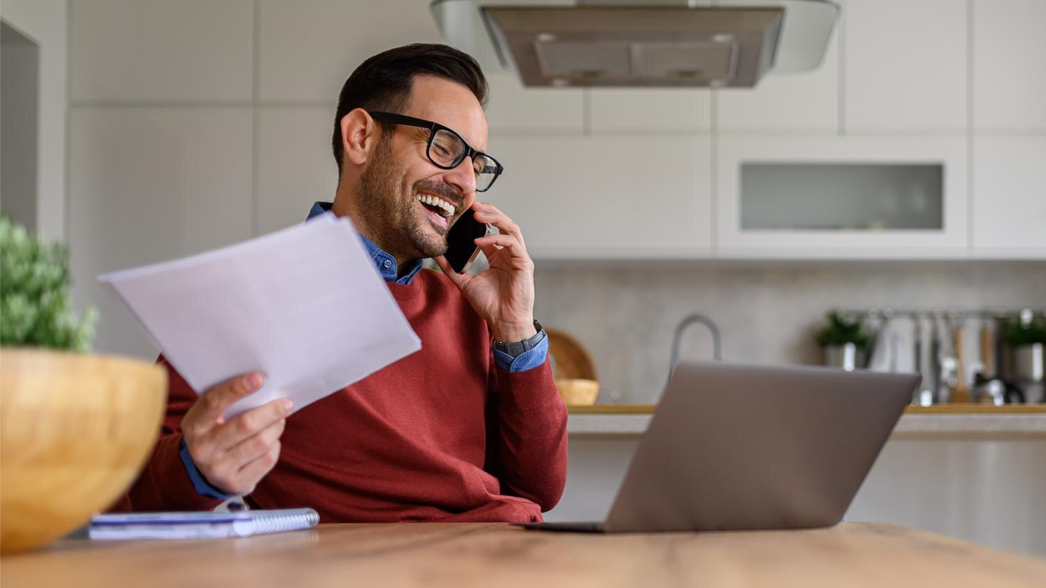 A man in glasses and a red sweater laughs while talking on the phone, holding printed documents in front of a laptop in a modern kitchen. This casual work-from-home moment aligns with VoIP call recording and Teams meeting recording for flexible communication compliance.