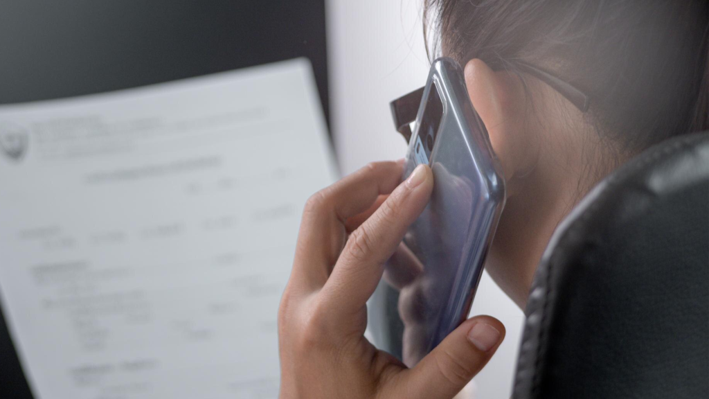 Close-up of a woman holding a smartphone to her ear during a conversation while viewing a document, representing a business scenario where Microsoft Teams phone call recording or Teams PSTN call recording might be used for compliance or accuracy.