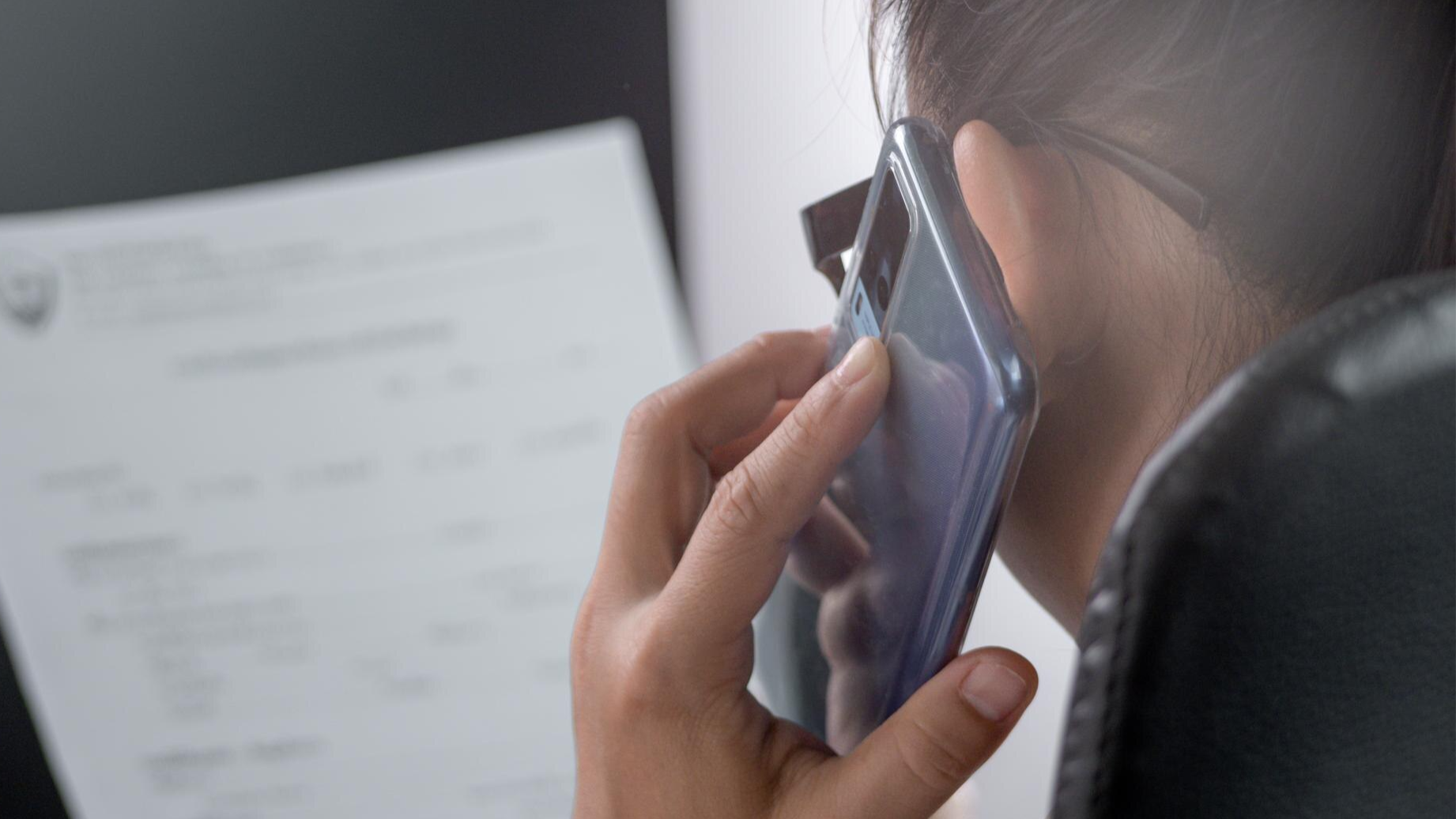 Close-up of a woman holding a smartphone to her ear during a conversation while viewing a document, representing a business scenario where Microsoft Teams phone call recording or Teams PSTN call recording might be used for compliance or accuracy.