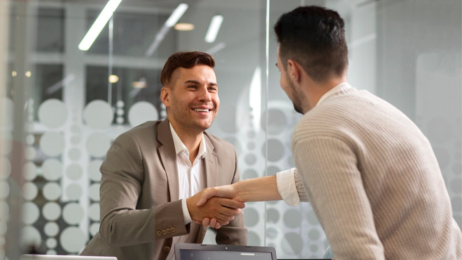 Two businessmen shaking hands across a desk in a modern office setting, suggesting the closure of a successful conversation or deal possibly held over a Microsoft Teams phone call with recording enabled for compliance documentation.