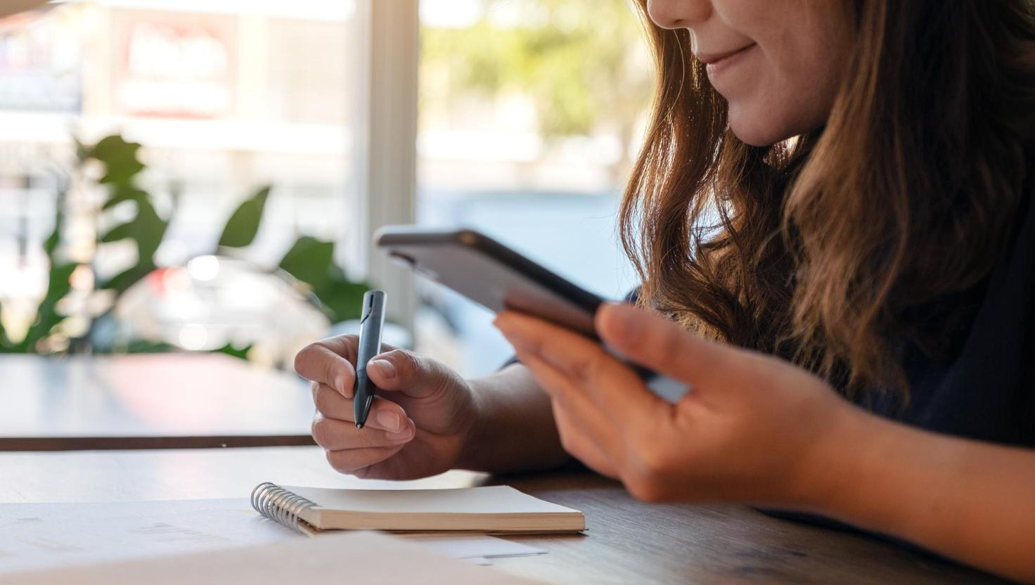 Close-up of a woman holding a smartphone and pen while writing in a notebook at a sunny desk. This scene can illustrate VoIP call recording and Microsoft Teams compliance solutions, especially for documenting key takeaways from Teams meeting recordings.
