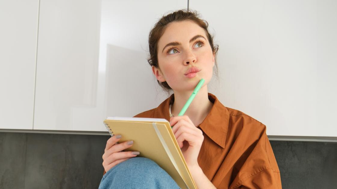 Young woman holding a notebook and pen while thoughtfully looking upward, possibly brainstorming or taking notes during a Teams call or reflecting on a previously recorded Microsoft Teams phone call.