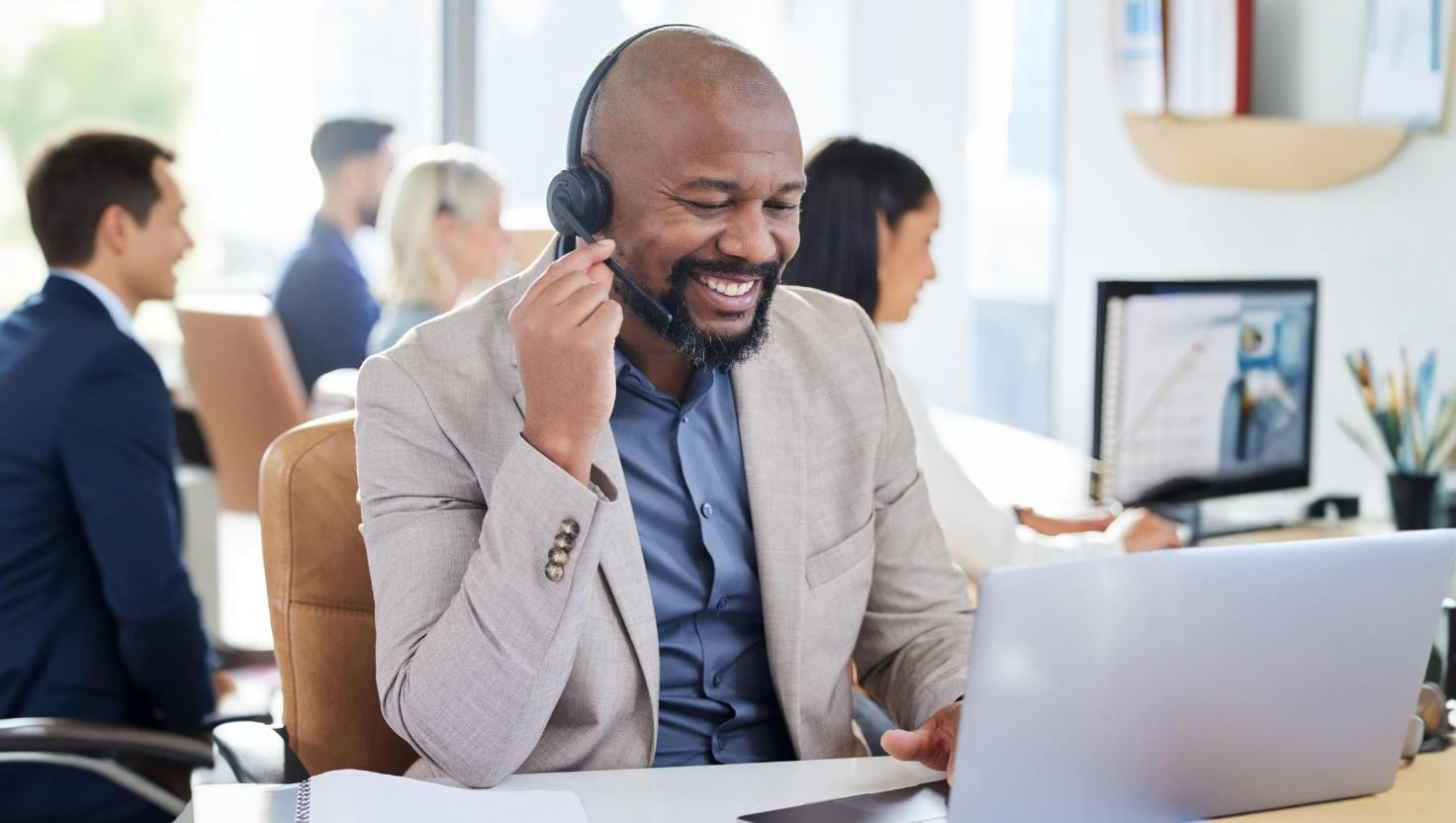 Smiling businessman in a headset conducting a virtual meeting on a laptop in an office, representing a professional environment where Teams call recordings or Microsoft Teams phone call recording features are commonly used.