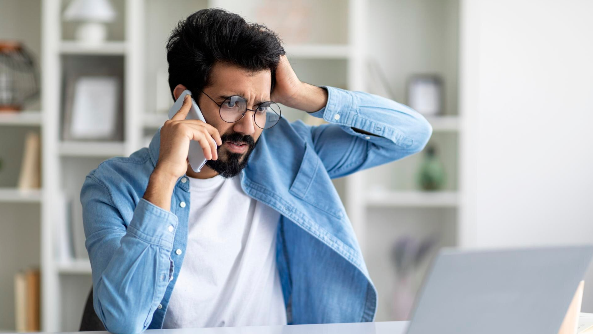 A concerned man speaks on the phone while looking at his laptop, with one hand on his head. This scenario may depict issues related to missing VoIP call recording or the need for reliable Teams call recording for compliance purposes.