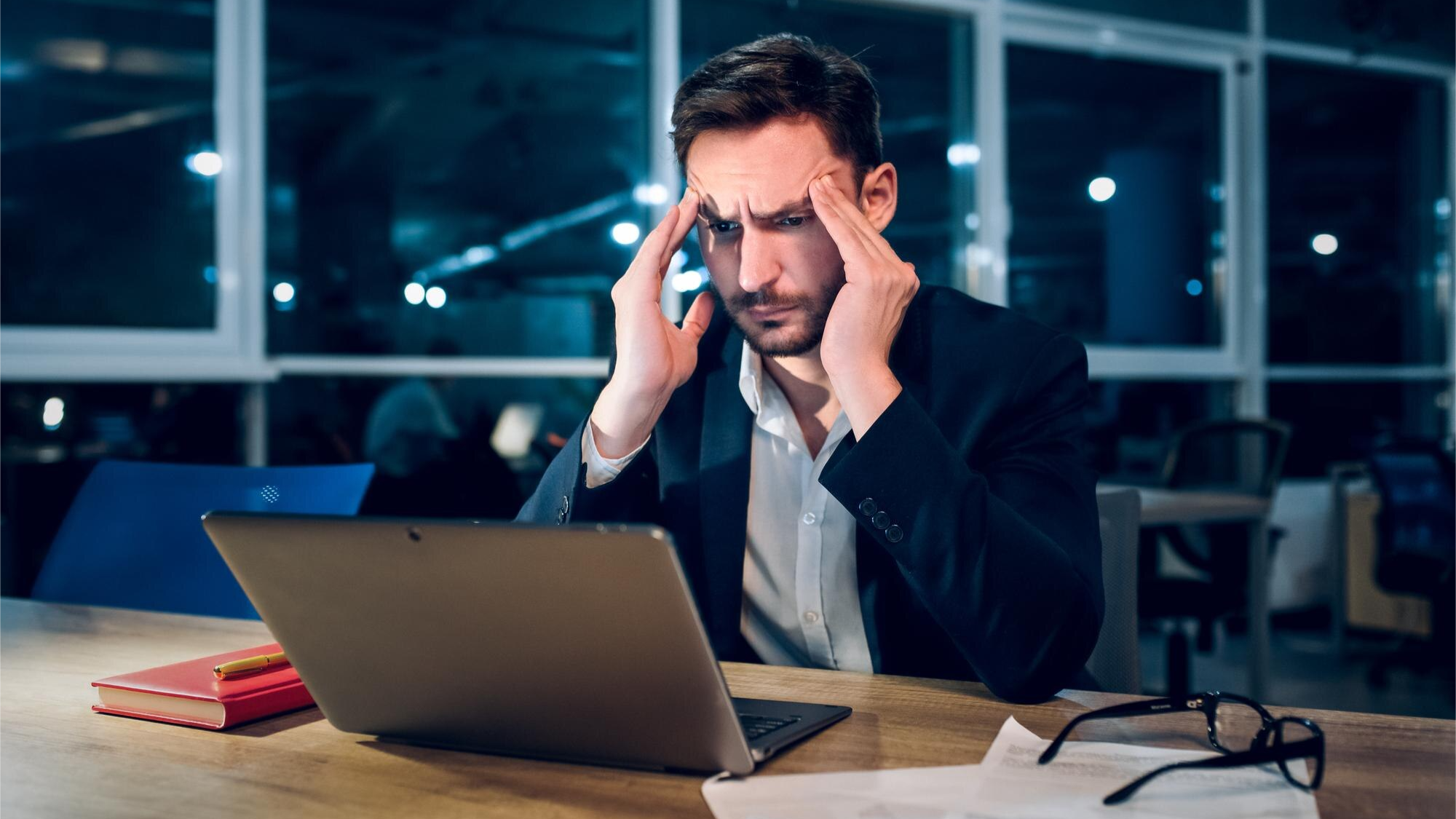 Stressed businessman in an office at night holding his head while looking at a laptop, possibly concerned about compliance or technical issues related to recording a Teams call or a Microsoft Teams phone recording.