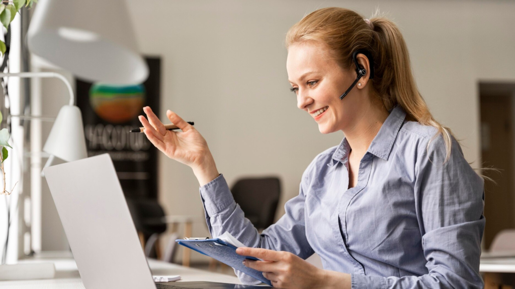 A smiling woman with a headset gestures during a video call, holding a clipboard in front of her laptop. This scene illustrates a professional using Microsoft Teams compliance recording or VoIP call recording as part of a cloud-based communication solution.