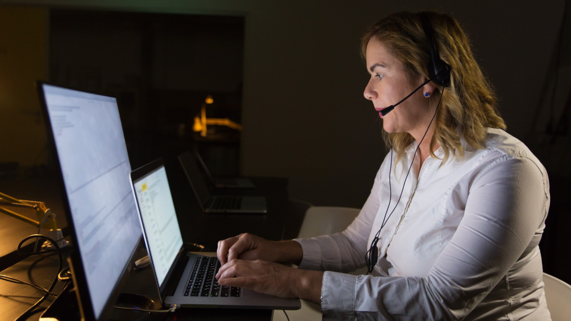 A woman in a headset works late at night, focused on her laptop and external monitors in a dimly lit room. This image reflects the importance of compliance call recording and Microsoft Teams call recording during extended work hours or global operations.