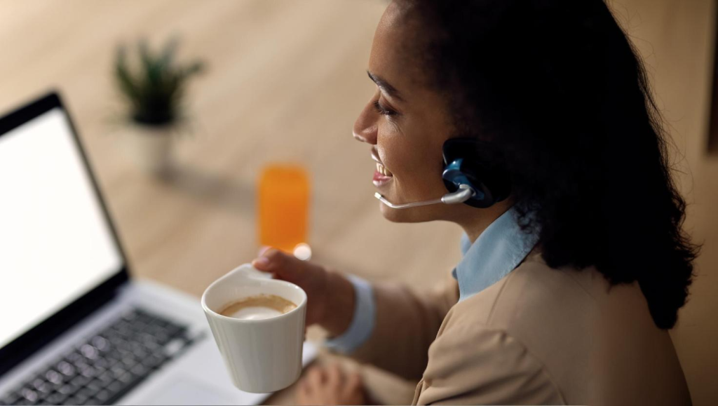 A smiling woman wearing a headset holds a coffee mug while participating in a video call on her laptop. This image represents Microsoft Teams Call Recording in a remote work setting, supporting compliance call recording for professional communication.