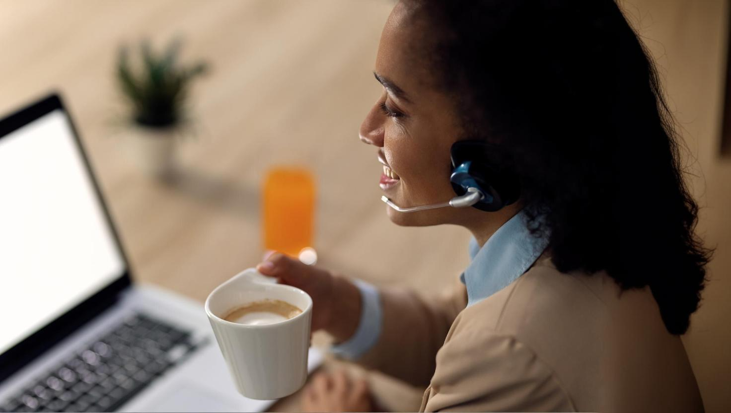 A smiling woman wearing a headset holds a coffee mug while participating in a video call on her laptop. This image represents Microsoft Teams Call Recording in a remote work setting, supporting compliance call recording for professional communication.