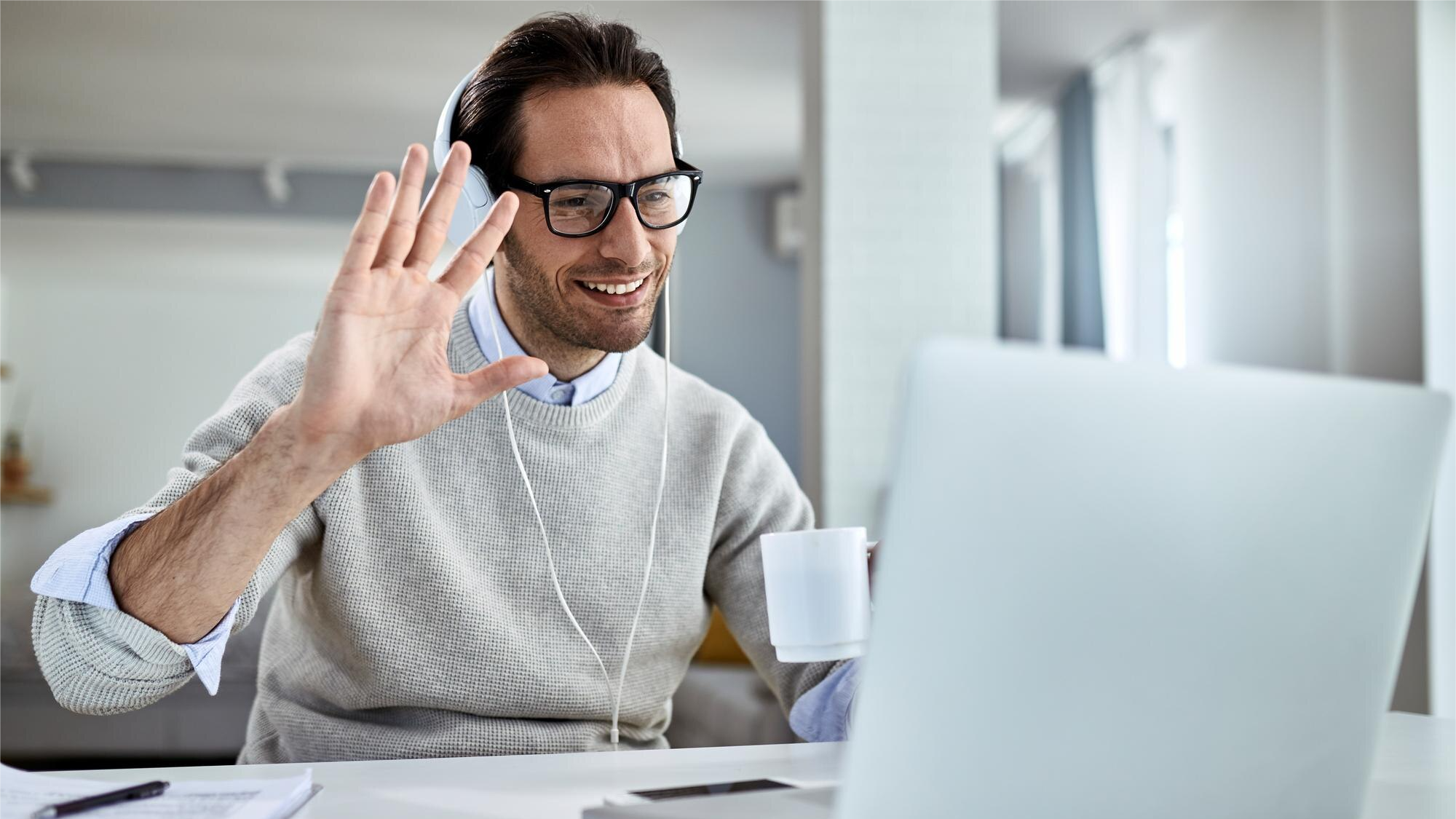 Man wearing glasses and headphones smiling and waving during a video call while holding a coffee mug, suggesting a friendly virtual meeting on Microsoft Teams, where recording the Teams call might be in use for future reference.
