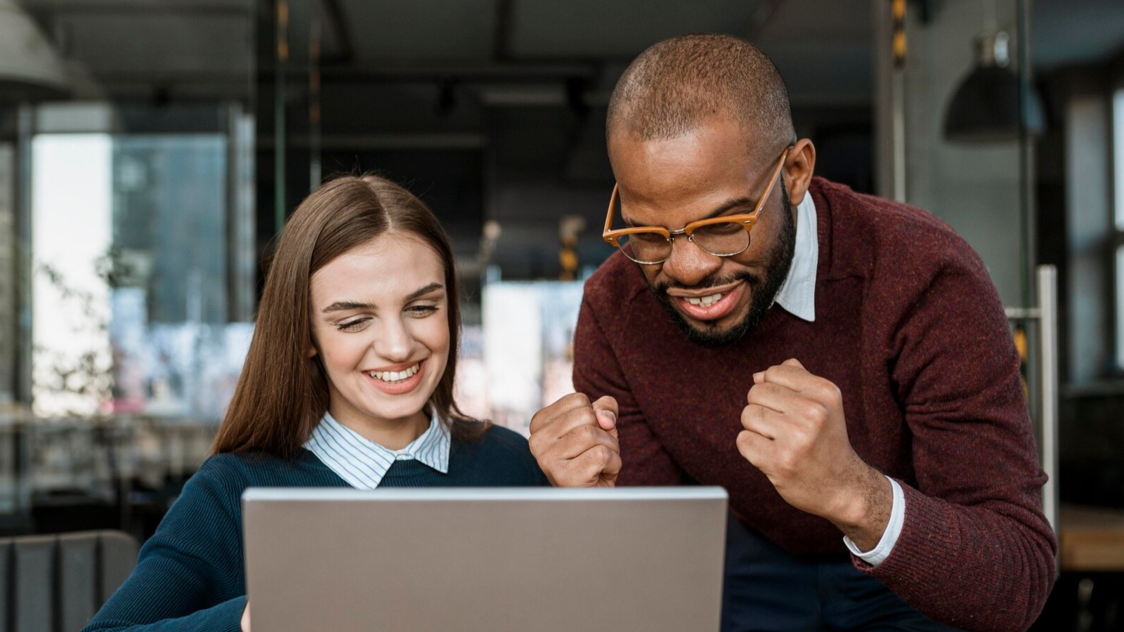 A woman is sitting at a laptop smiling while a man is standing beside her with both fists clenched in excitement.